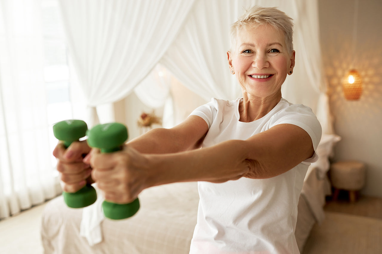 50+ year old smiling woman working out with dumbbells in her bedroom depicting healthy longevity